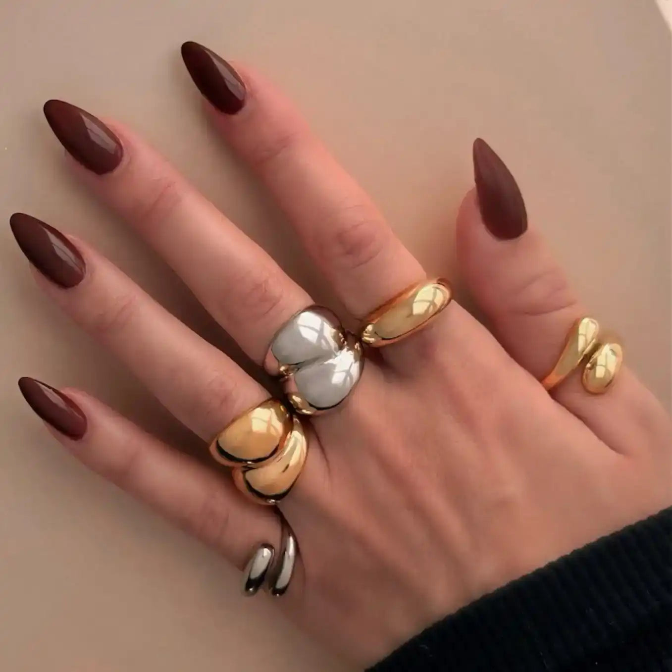 Hand wearing multiple gold and pearl statement rings with dark burgundy nail polish.